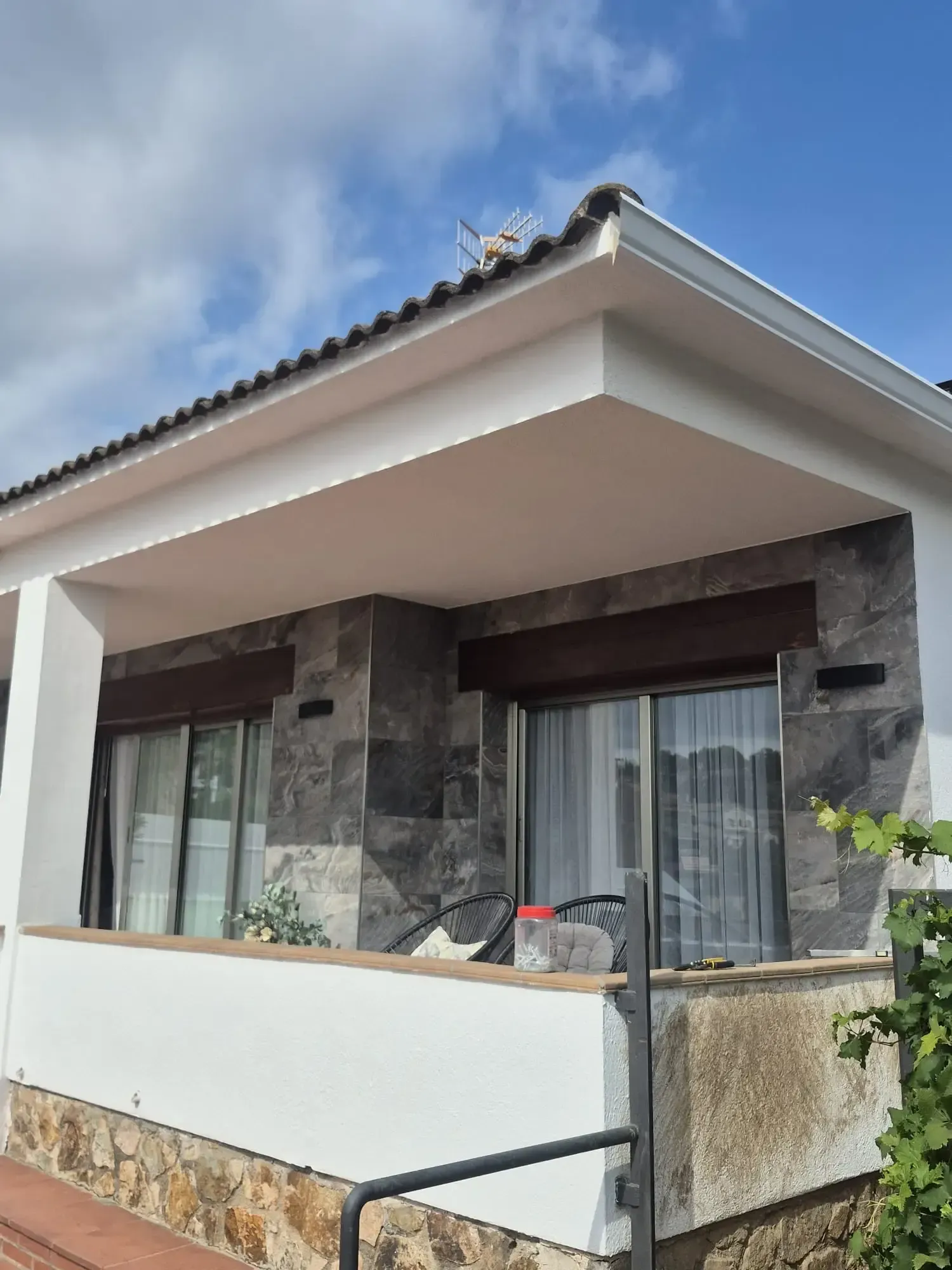 Porche de una casa moderna con muros de piedra, barandilla blanca y tejado de tejas bajo un cielo despejado