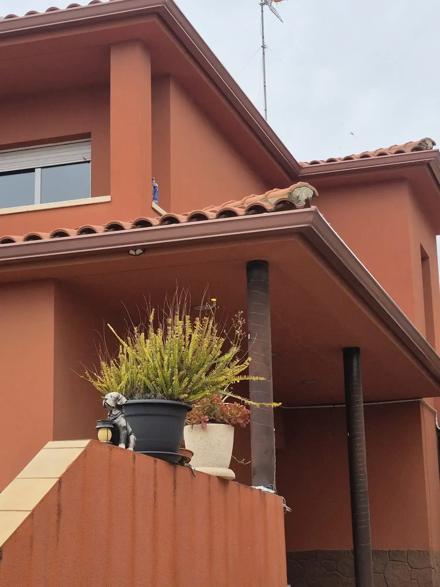 Exterior de una casa de terracota con tejado de tejas, balcón y plantas en macetas sobre un muro de ladrillo