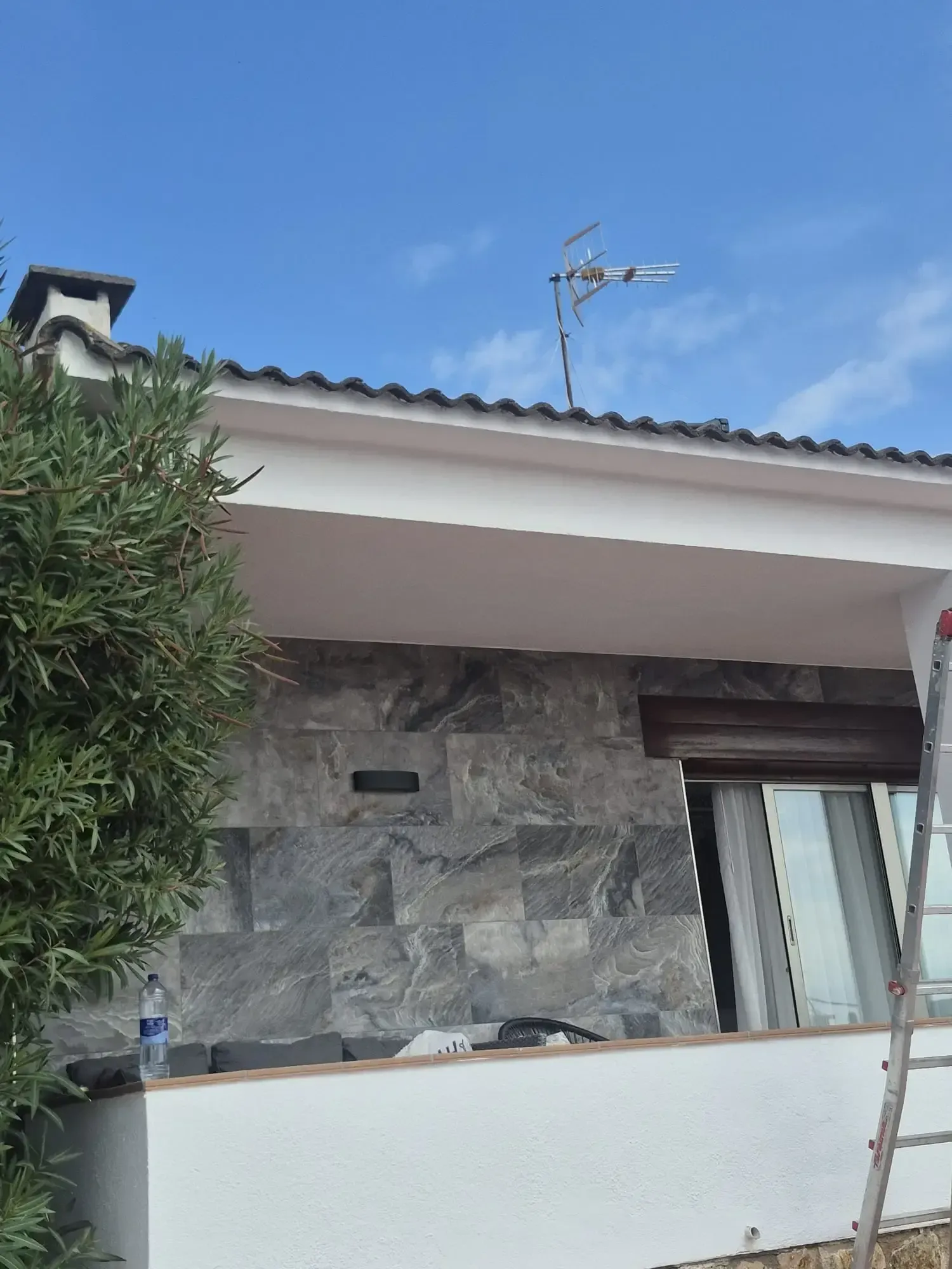 Balcón con pared de piedra, barandilla blanca, cielo azul y una escalera apoyada en el lado derecho