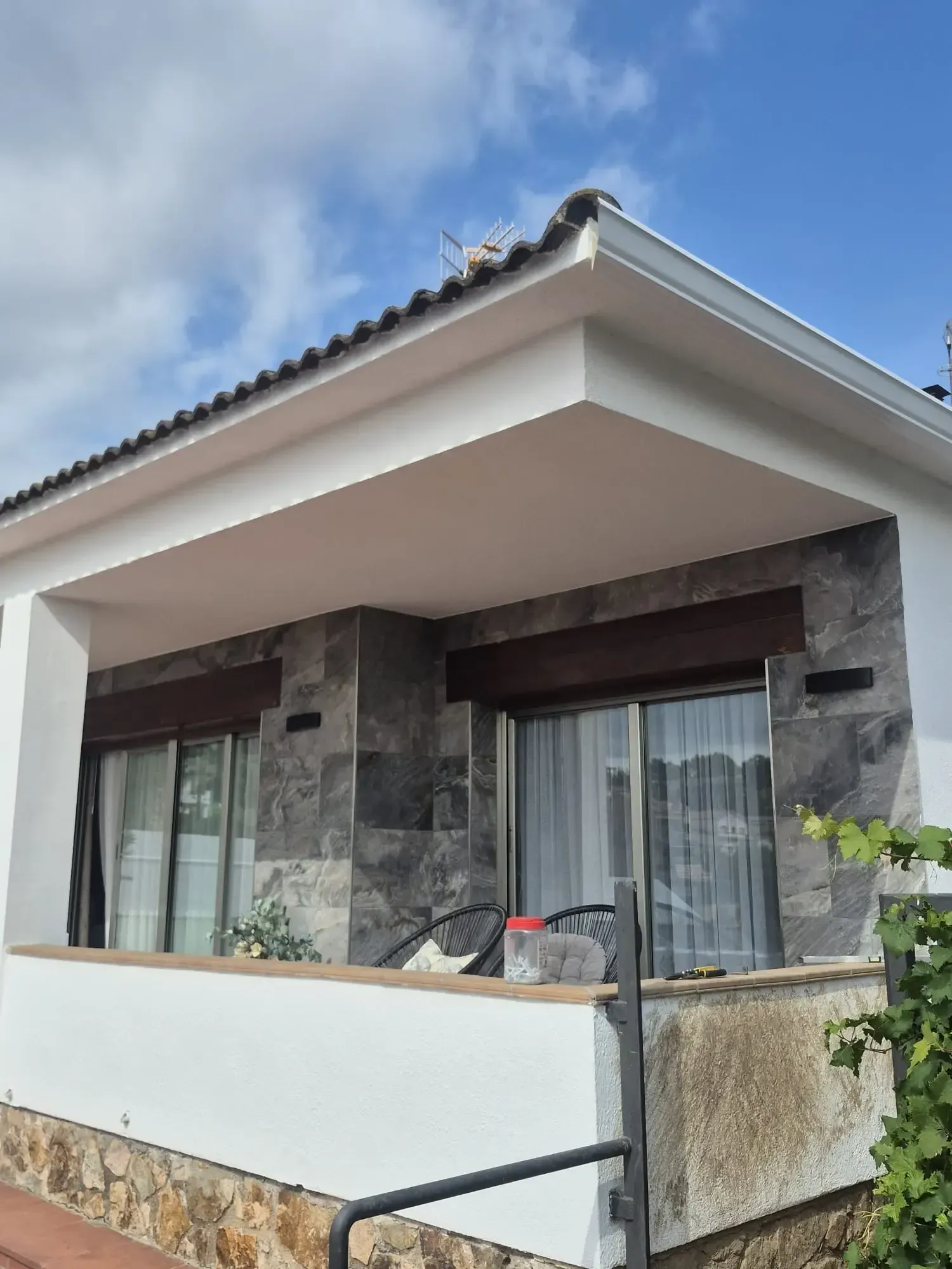 Exterior de una casa moderna con fachada de piedra, pared blanca en el balcón y grandes ventanales bajo un tejado de tejas