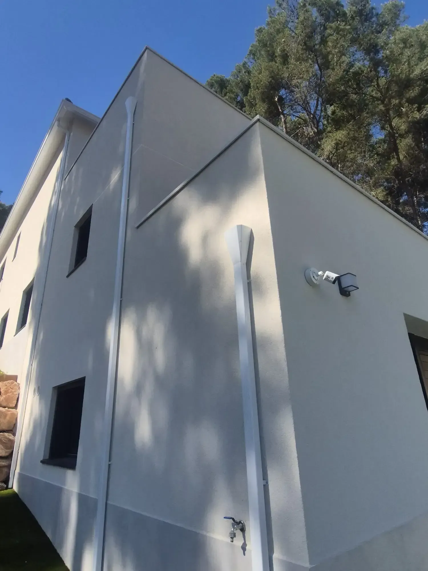 Exterior de una casa de estuco blanco con una ventana pequeña, una lámpara en la pared y árboles que proyectan sombras