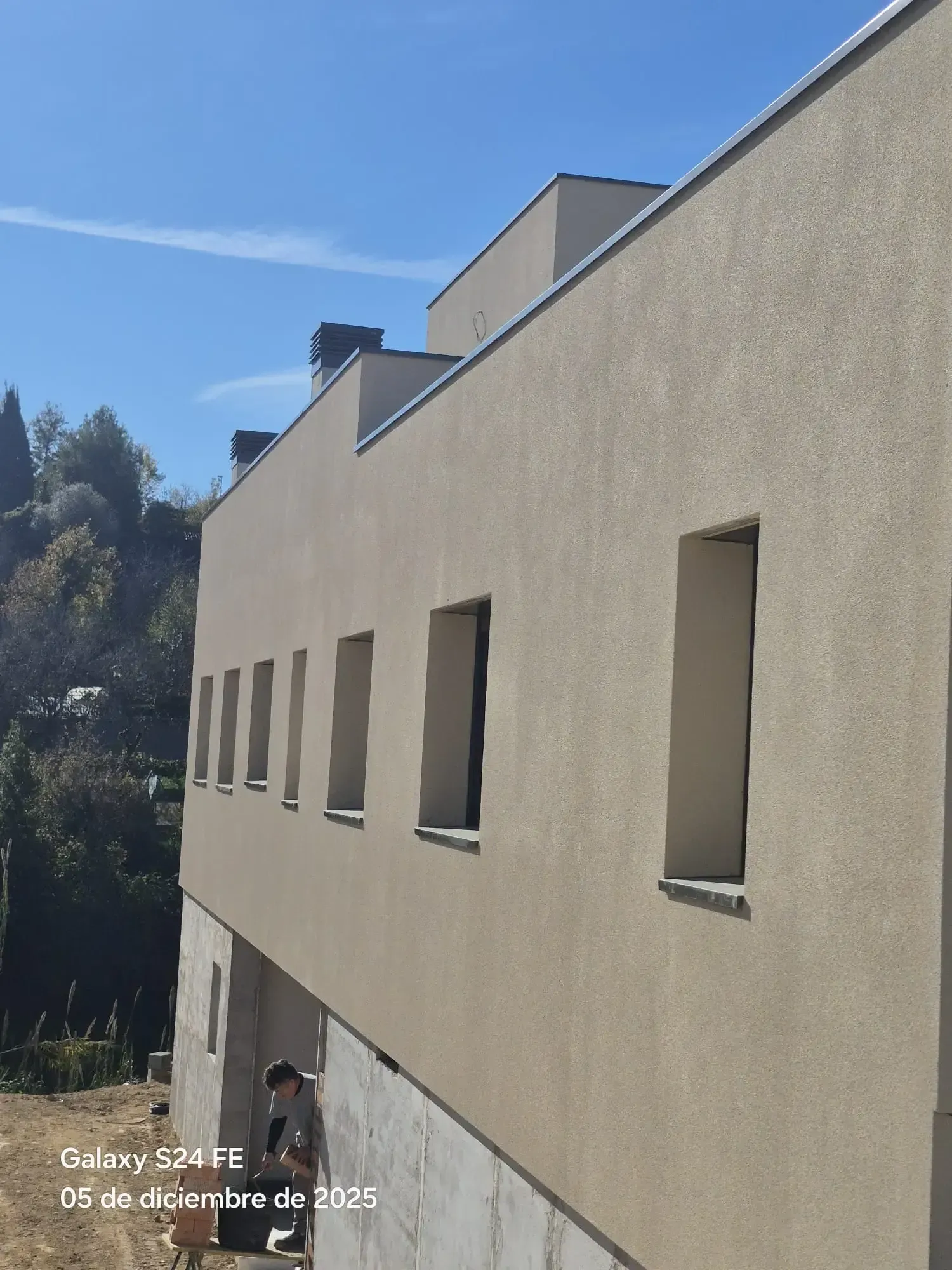 Fachada de un edificio beige con ventanas pequeñas bajo un cielo azul despejado, vista en perspectiva.