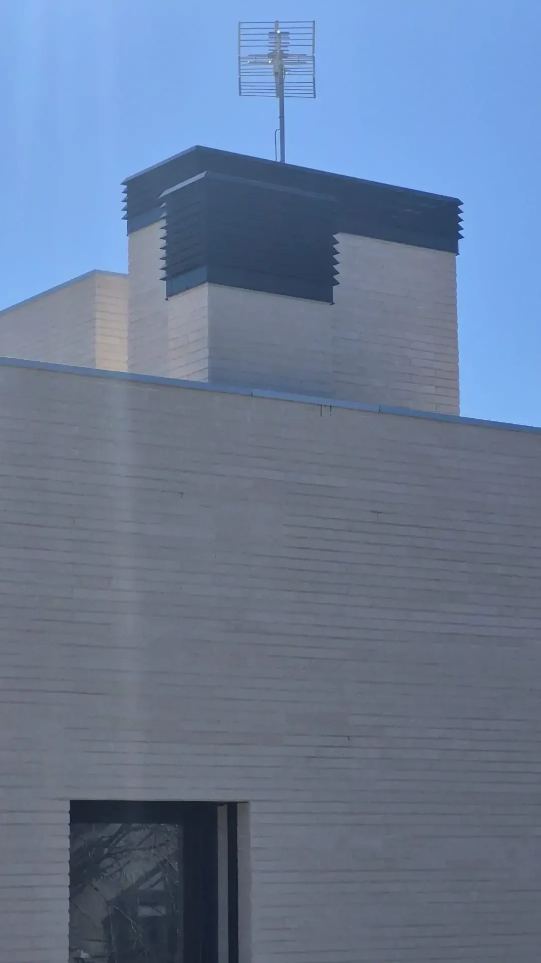Fachada de edificio blanco con una antena en el tejado y una rejilla de ventilación negra bajo un cielo azul despejado