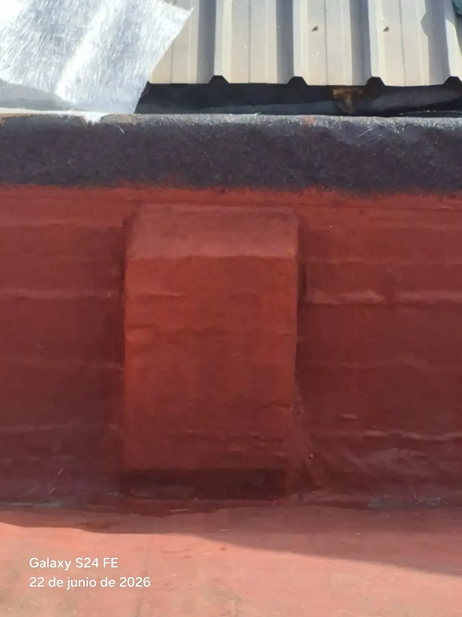 Pared roja con una puerta centrada bajo un tejado de chapa ondulada, iluminada por el sol.