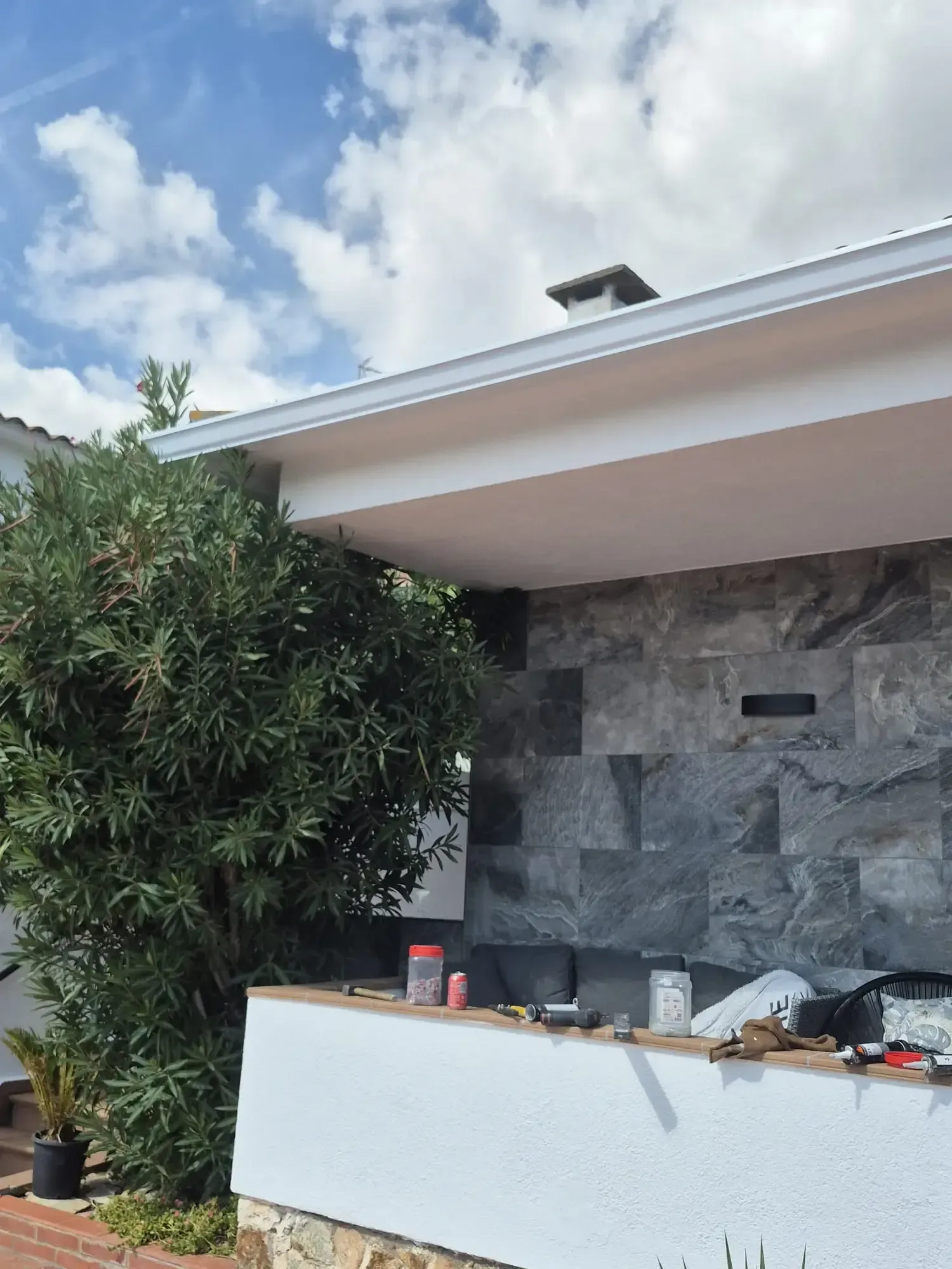 Zona de bar al aire libre con una barra blanca, un muro de piedra, plantas tropicales y un cielo azul