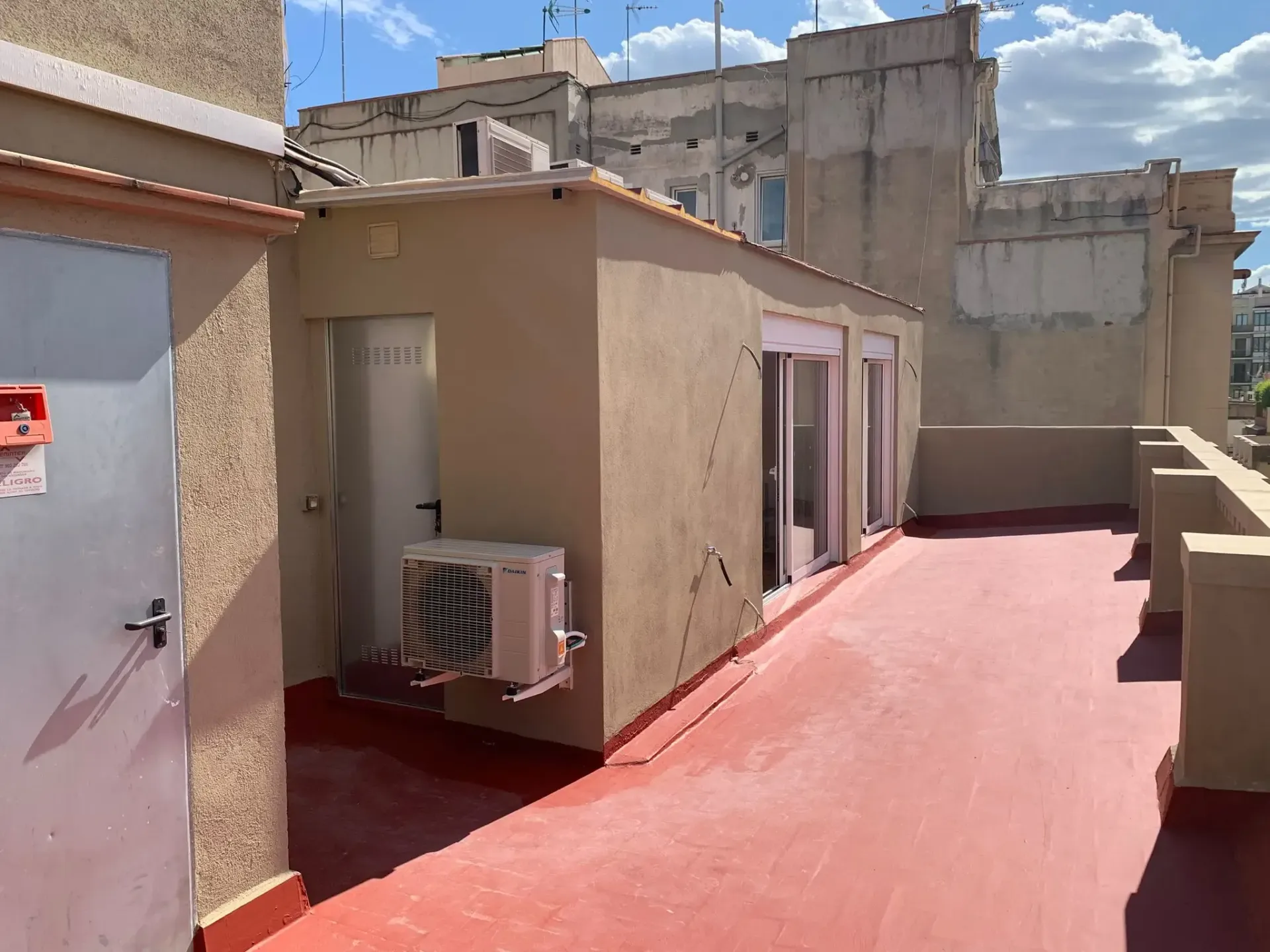 Terraza en la azotea con suelo rojo, cuarto de servicio beige, aire acondicionado de pared y edificios de apartamentos alrededor