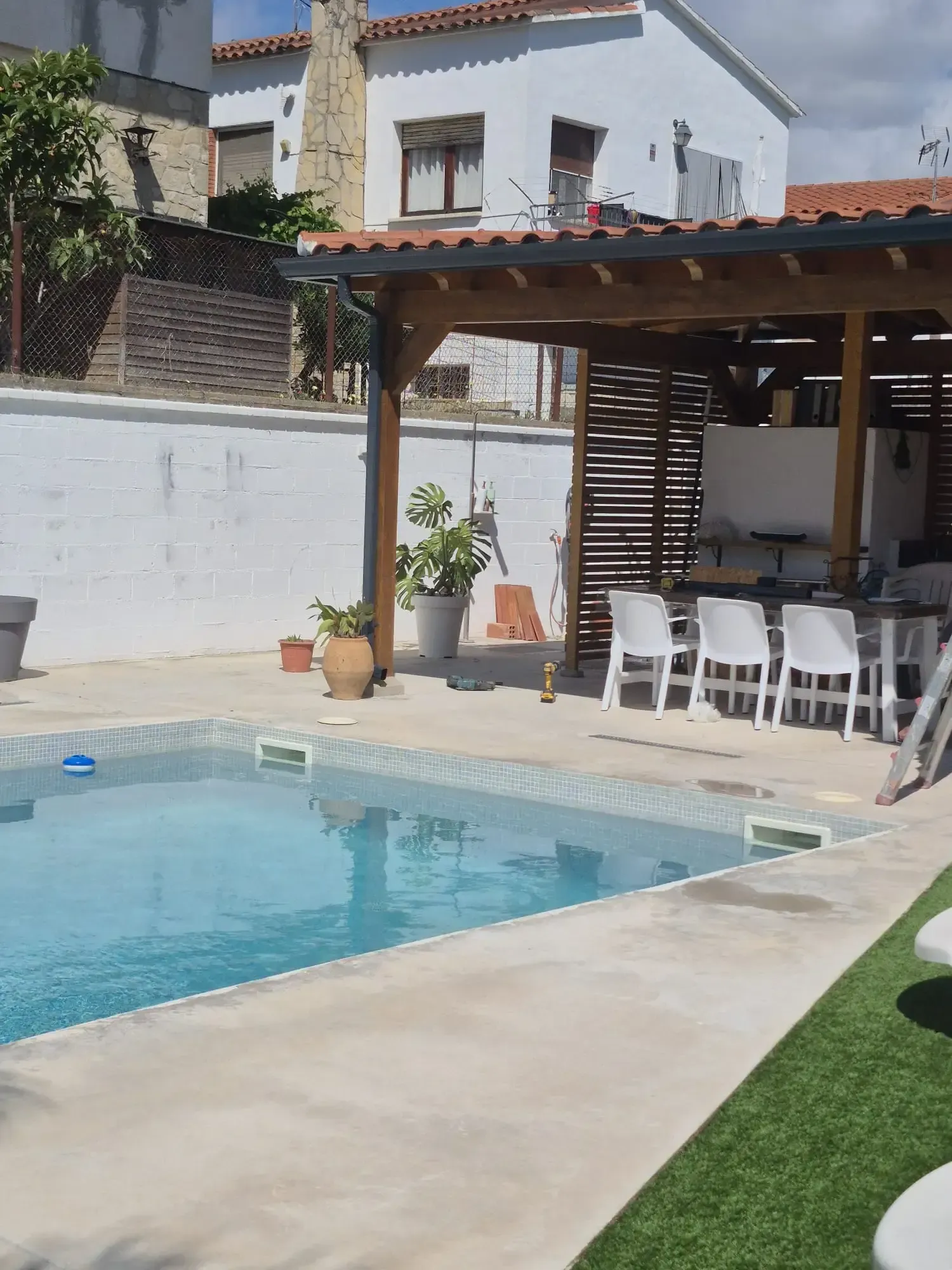 Piscina en el patio trasero junto a un porche cubierto con sillas blancas, plantas en macetas y una franja de césped