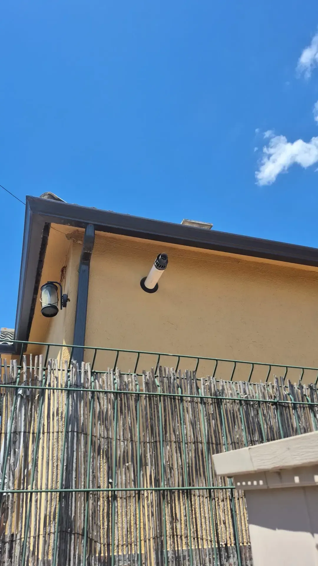 Edificio de estuco color arena con dos apliques en la pared, detrás de una cerca de bambú y bajo un cielo azul