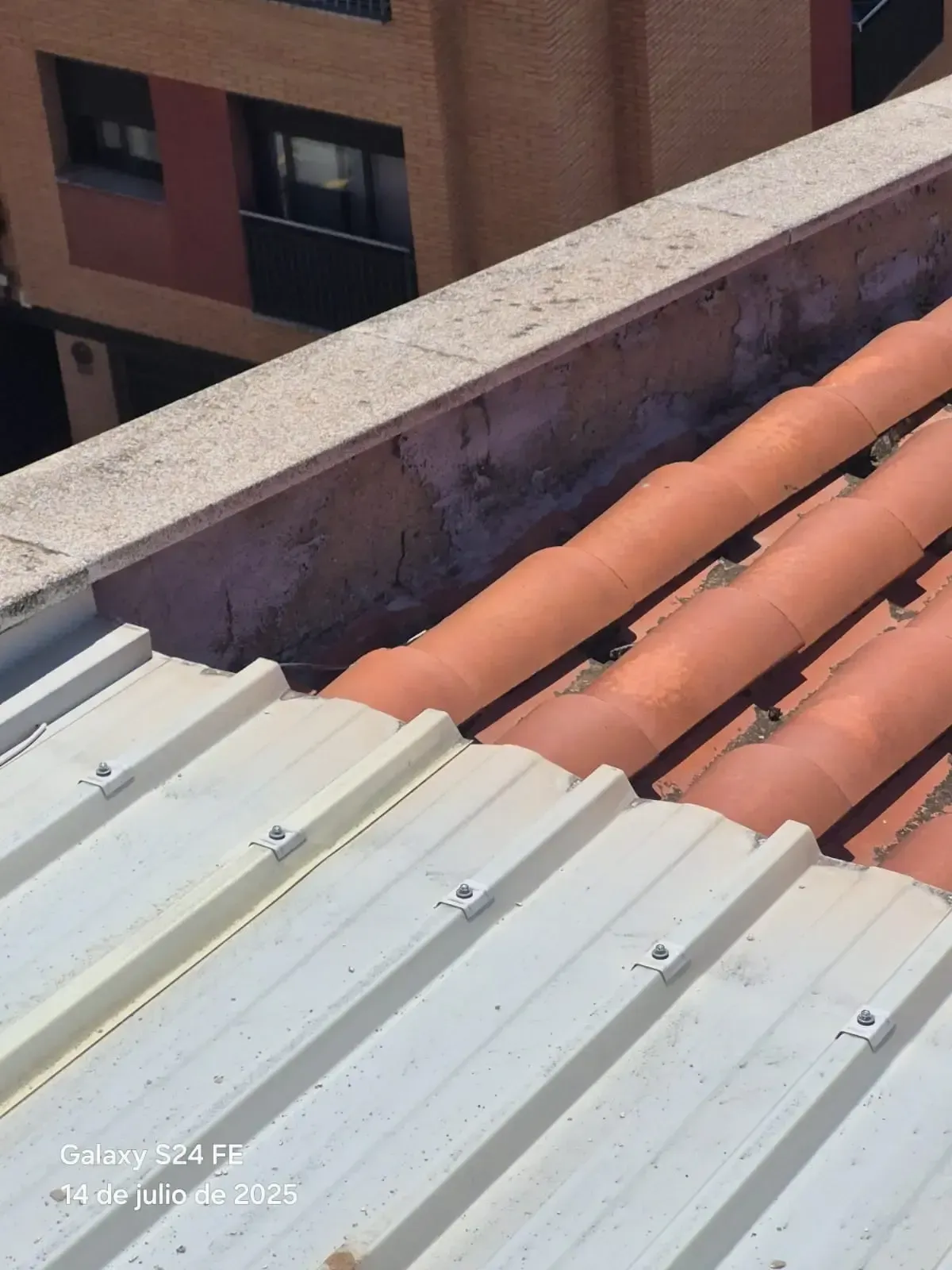 Tejado con chapa ondulada blanca y tejas de barro anaranjadas junto a un parapeto de hormigón, con vistas a edificios de apartamentos