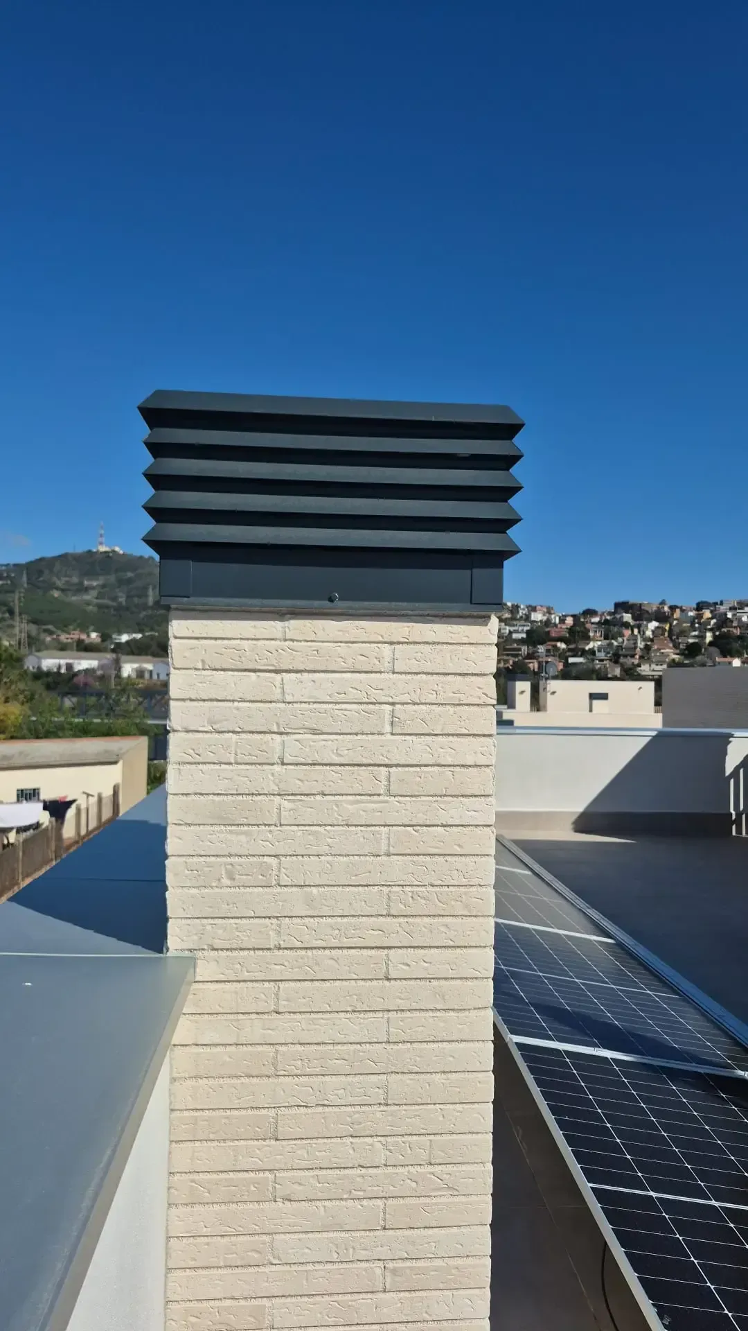 Chimenea de ventilación en la azotea, con listones oscuros sobre una columna de ladrillo y el cielo azul y el paisaje urbano de fondo