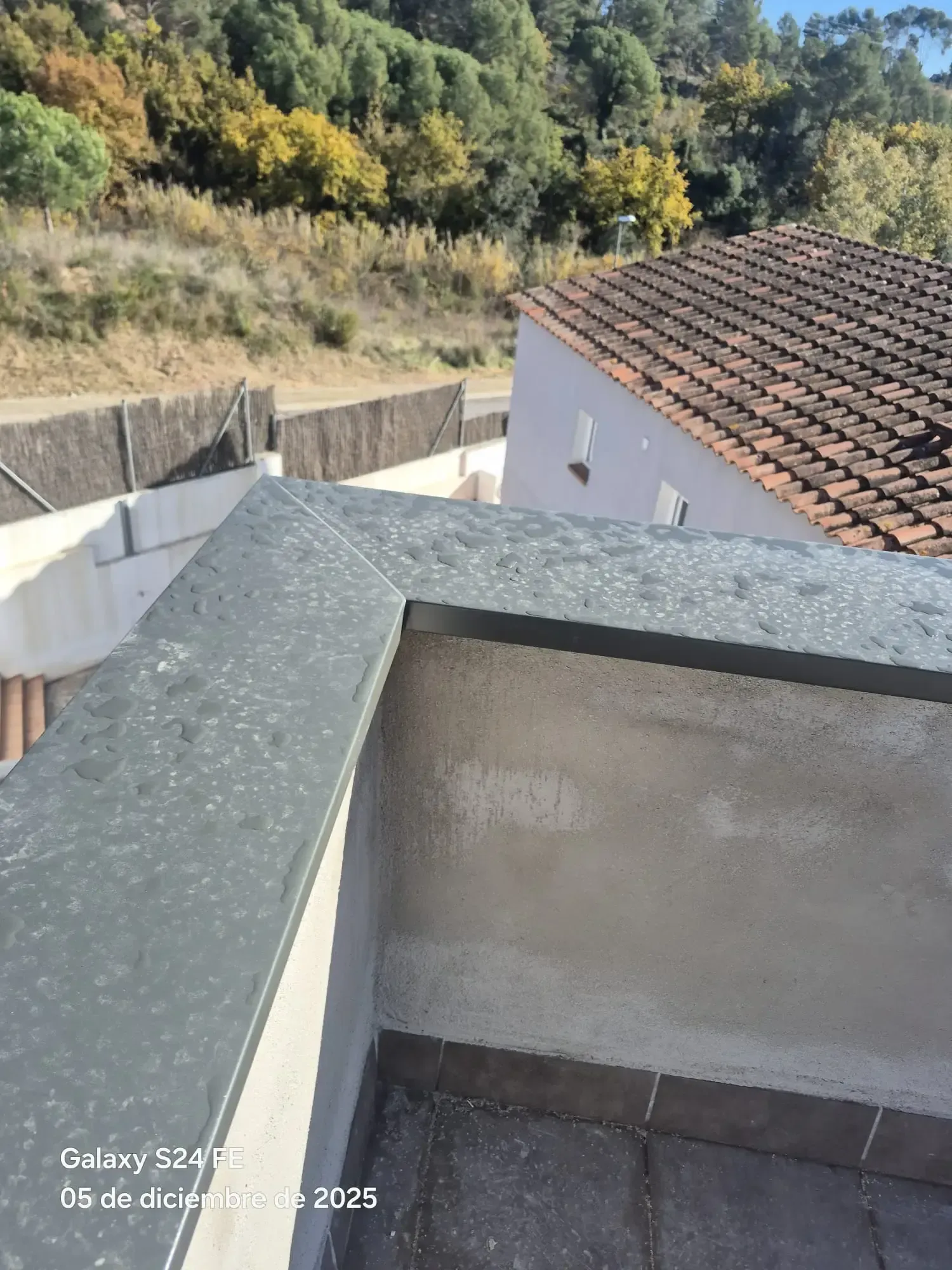 Terraza en la azotea con pretil de hormigón, con vistas a un tejado de tejas rojas y una ladera boscosa.