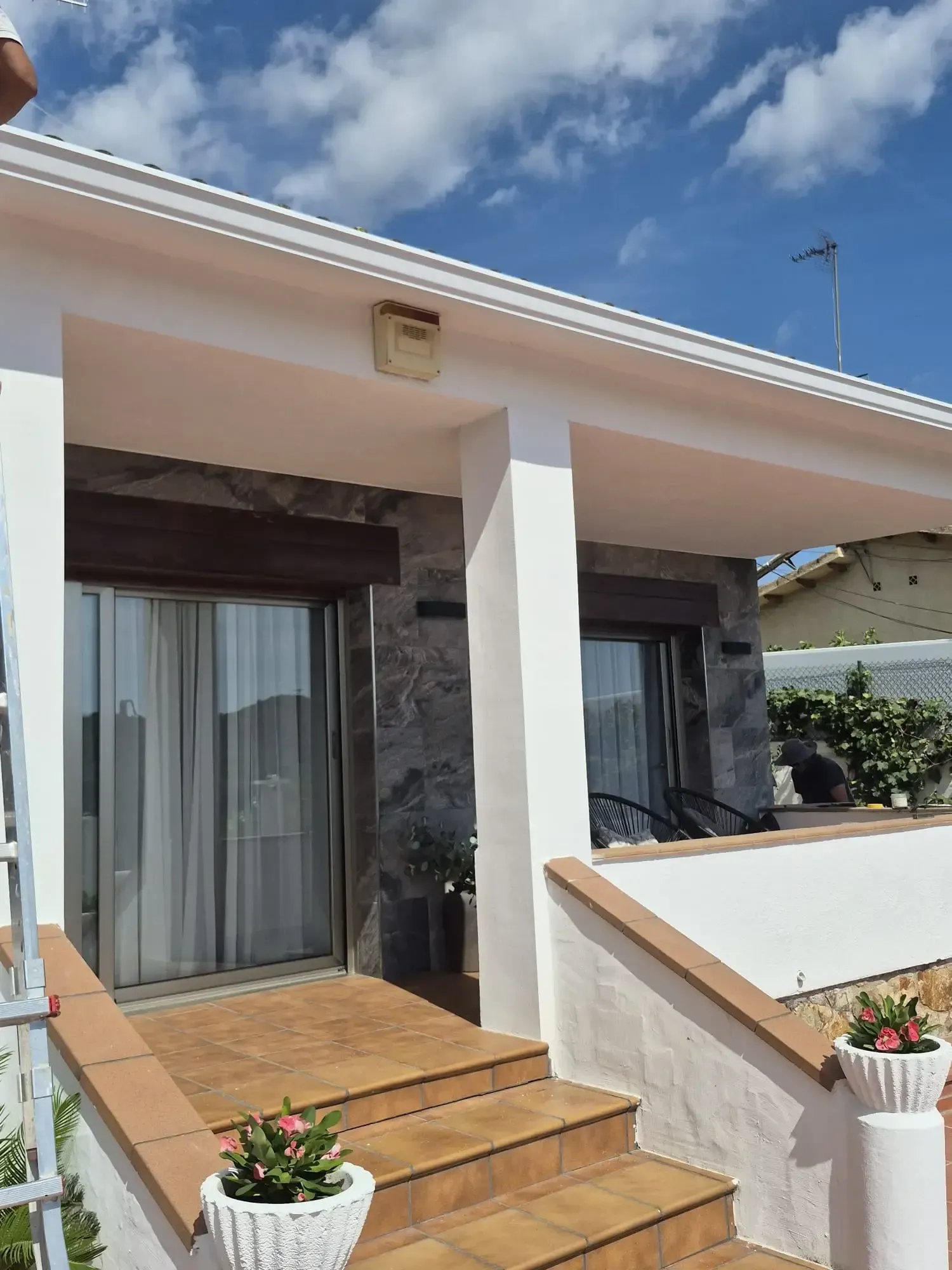 Entrada principal de una casa blanca con escalones color tostado, ventanas con marcos negros y plantas en macetas bajo un cielo azul