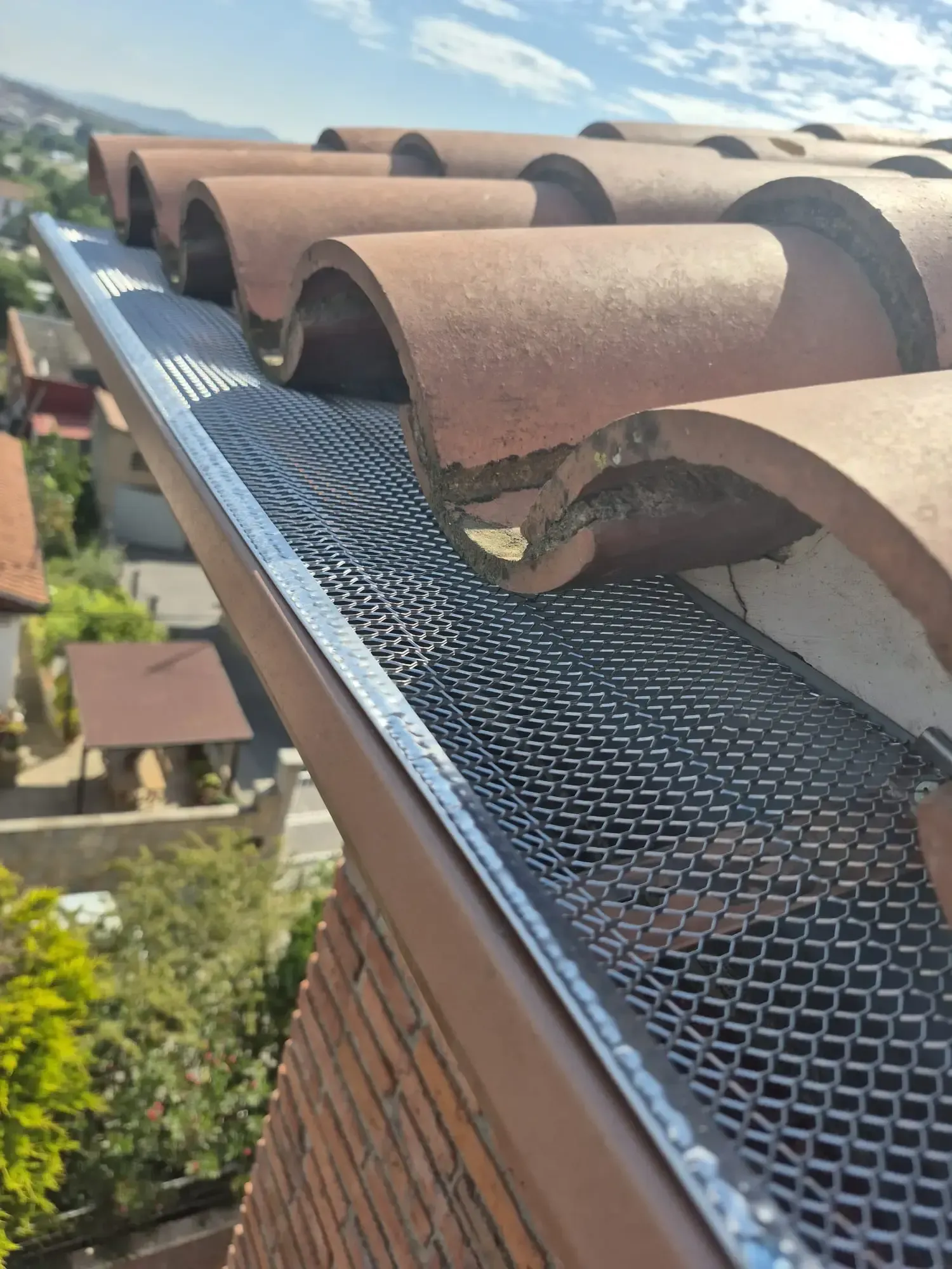 Borde del tejado con protector de canalón negro bajo tejas curvas de terracota, con vistas a un pueblo en la ladera de una colina