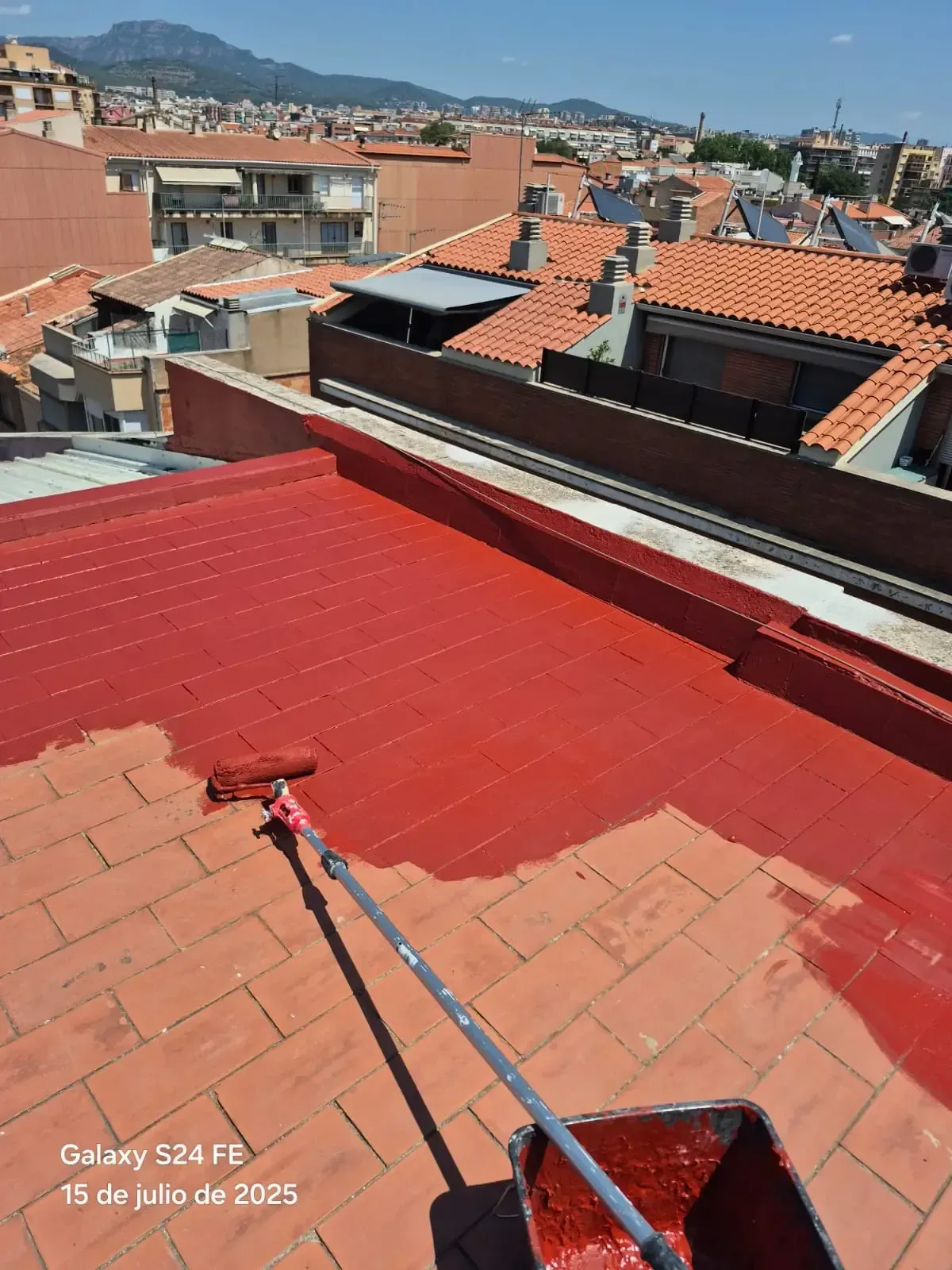 Pintura de azotea con revestimiento impermeabilizante rojo sobre tejas de terracota, con el perfil urbano de la ciudad al fondo.