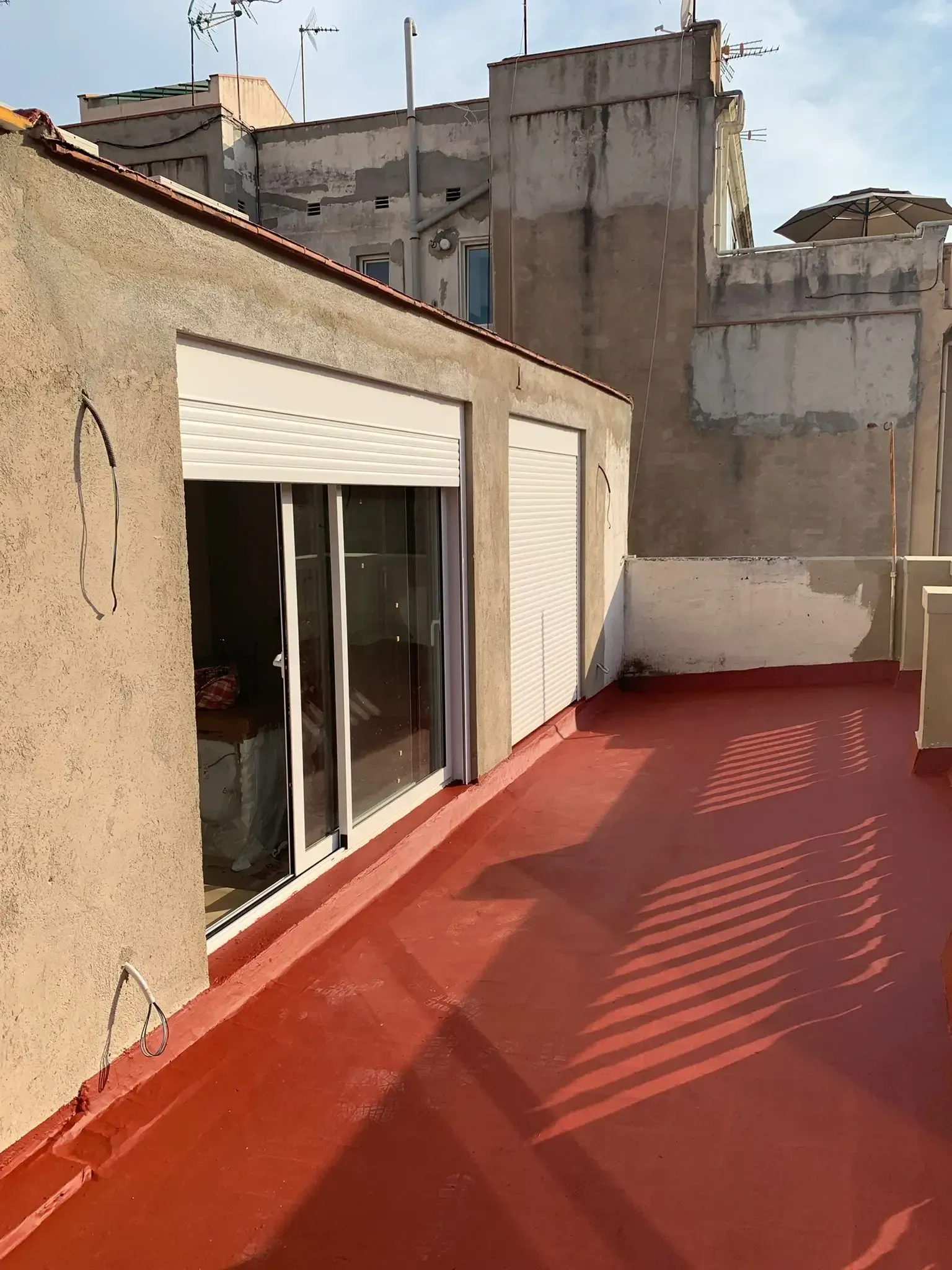 Terraza en la azotea con suelo rojo, puertas correderas de cristal y edificios de hormigón alrededor, bajo un sol radiante