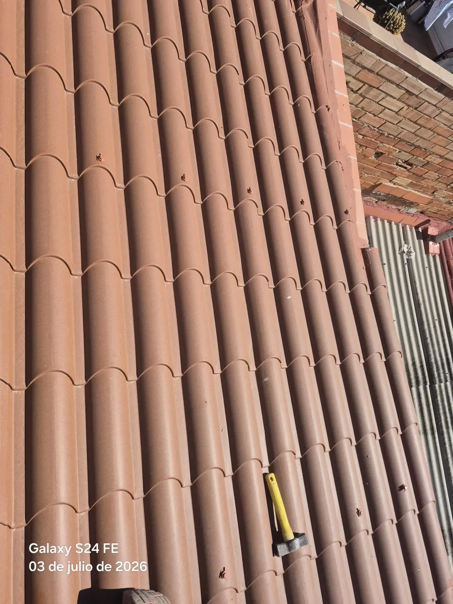 Tejas de terracota apiladas junto a una pared de ladrillo, con una herramienta amarilla colgada cerca de la parte inferior.