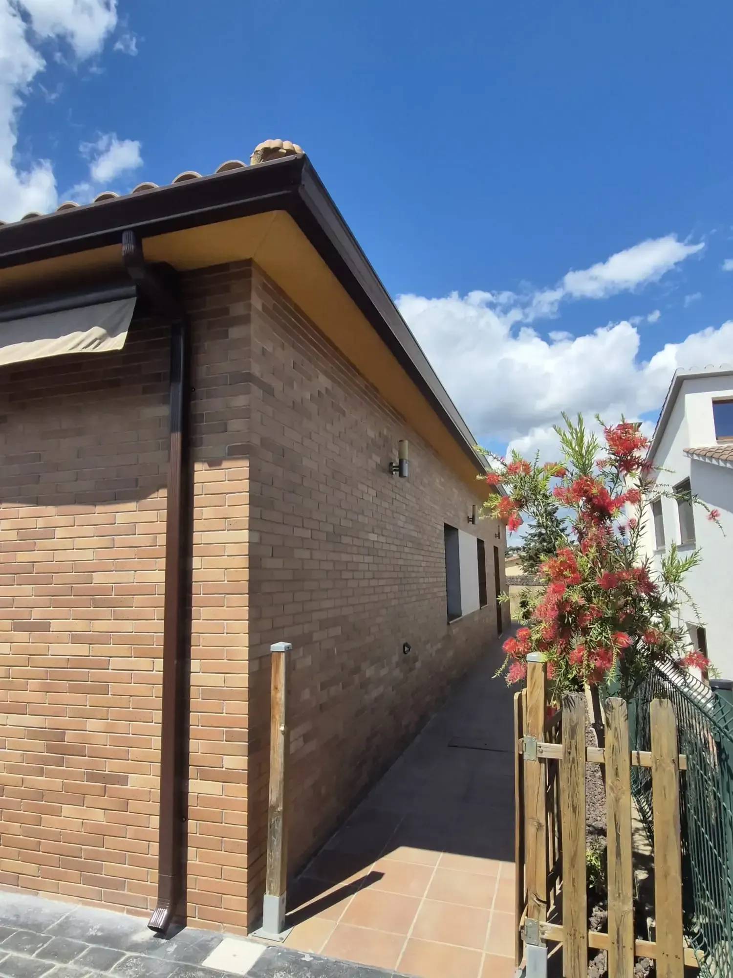 Pasillo lateral de una casa estrecha de ladrillo, con camino de baldosas, cerca de madera y cielo azul con nubes