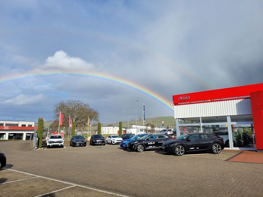 Über einem Autohaus ist ein Regenbogen zu sehen, davor parkende Autos.