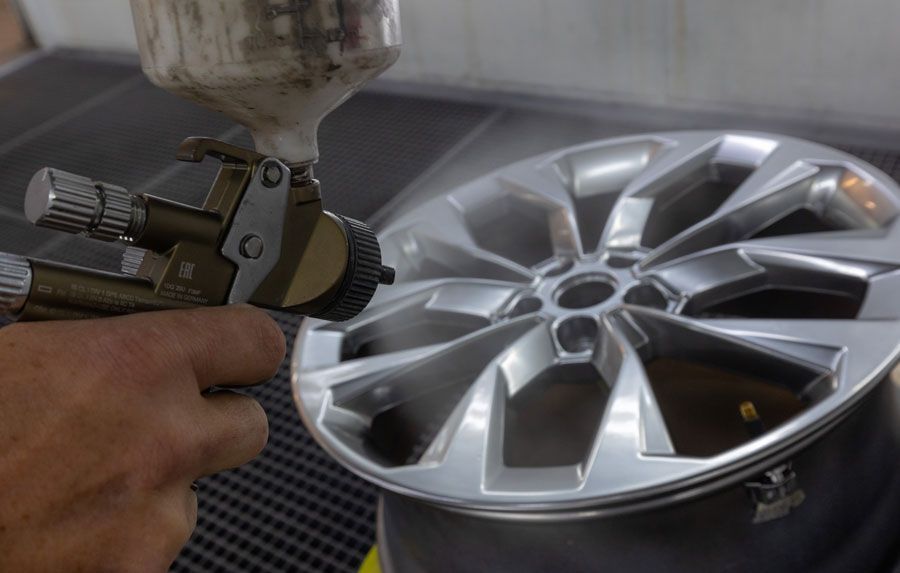 Hand, die in einer Werkstatt eine silberne Autofelge mit einem Farbsprühgerät lackiert.
