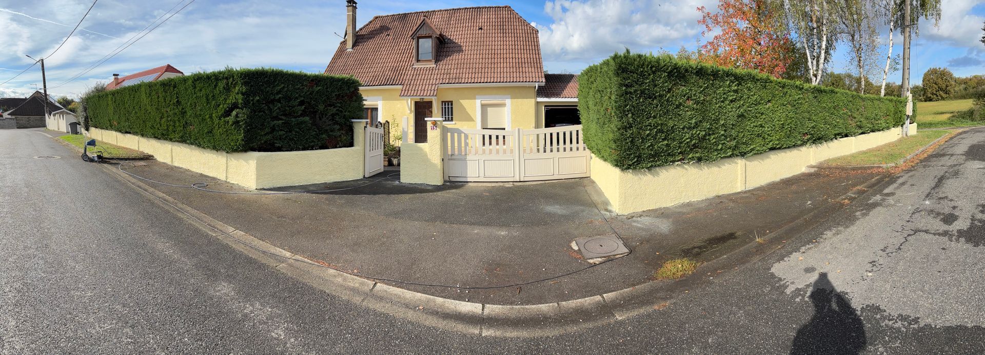 Une maison jaune au toit rouge, entourée de haies vertes et d'une rue.