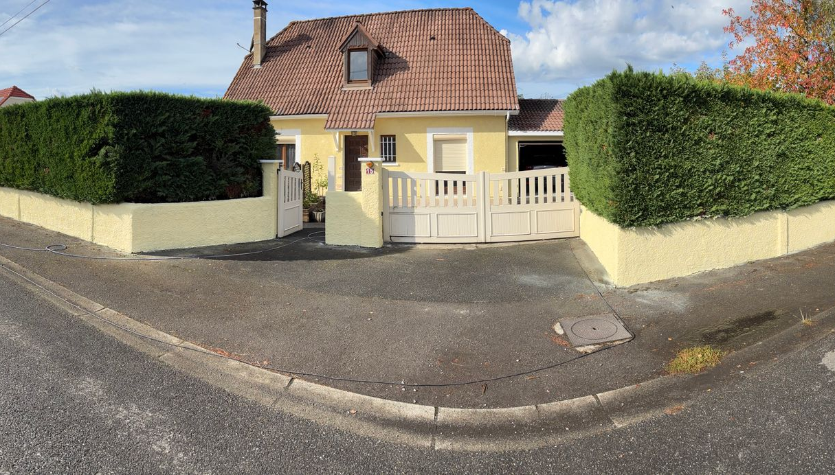 Maison jaune au toit rouge et aux haies vertes, vue de la rue.