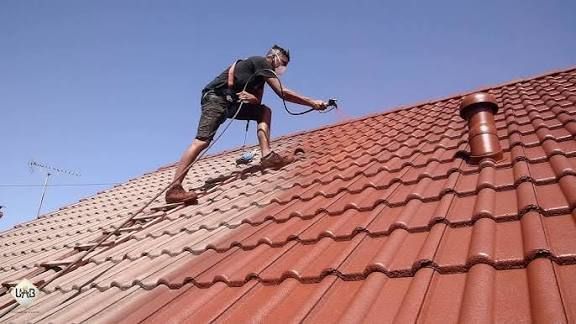 Une personne peint un toit de tuiles rouge, maintenue par un harnais. Ciel bleu clair en arrière-plan.
