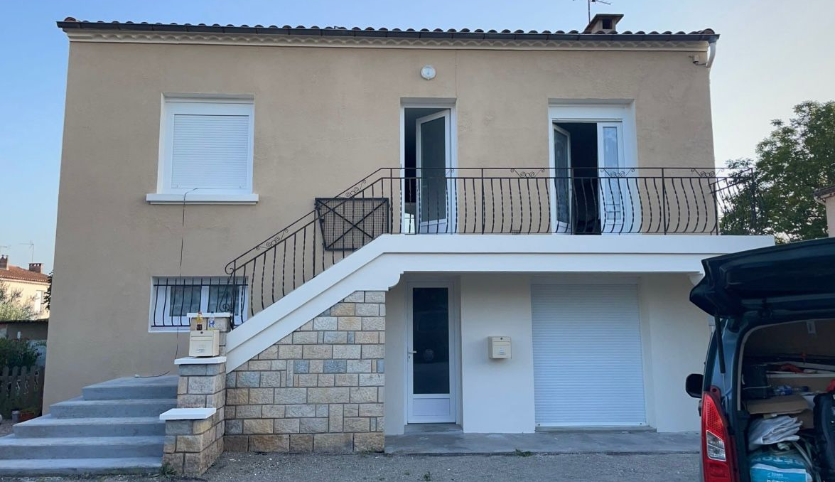 Maison à deux étages avec façade en stuc beige, boiseries blanches, balcon ouvert et escalier extérieur.