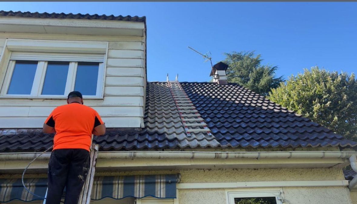 Une personne en chemise orange, perchée sur une échelle, nettoie un toit de tuiles par une journée ensoleillée.