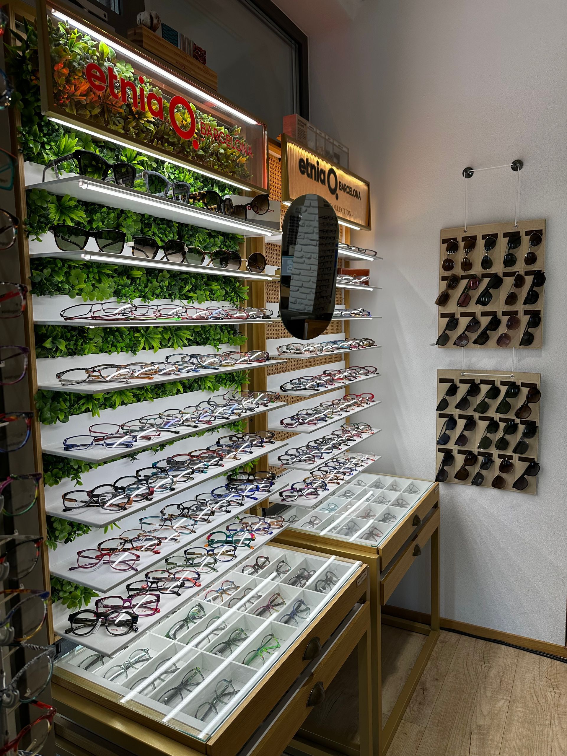 Das Interieur eines Optikergeschäfts mit Reihen von ausgestellten Brillengestellen, einem zentralen Bedientresen und grauen Stühlen.