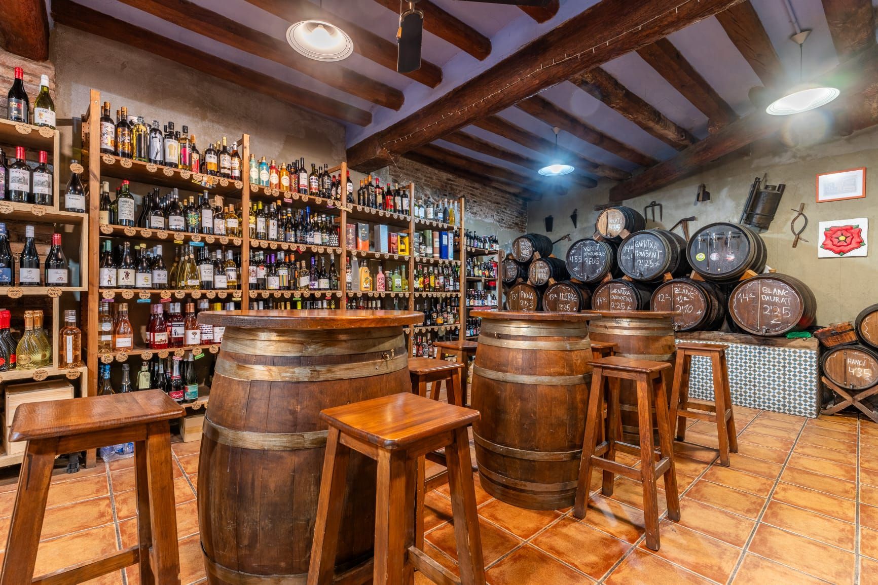 Una bodega rústica con numerosos barriles de madera, taburetes de bar y amplias estanterías repletas de botellas de vino.