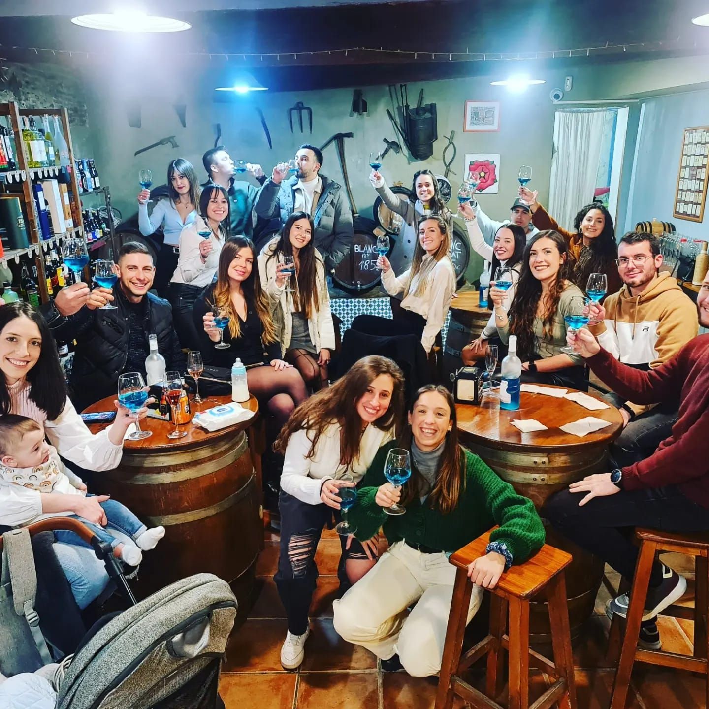 Un numeroso grupo de personas sonríe y brinda durante una celebración en una bodega rústica decorada con barriles.