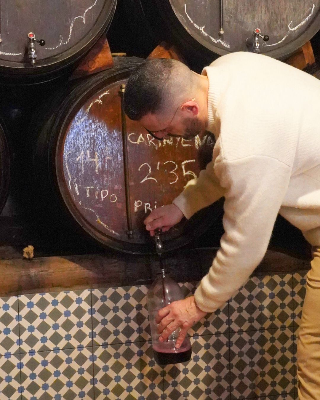 Una persona llena una botella de vidrio con vino tinto de un barril de madera en una bodega con paredes de azulejos.