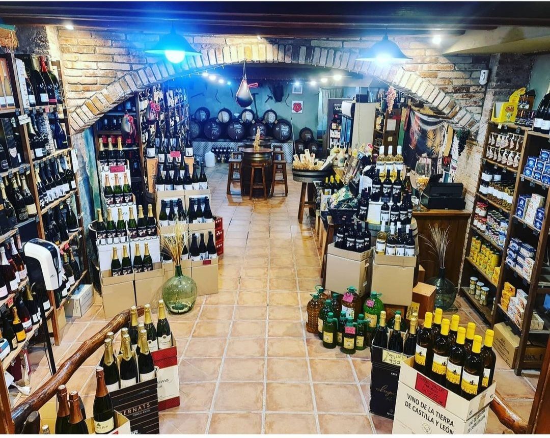 Una vinoteca con arcos de piedra, estanterías repletas de botellas y una mesa central con barriles al fondo.