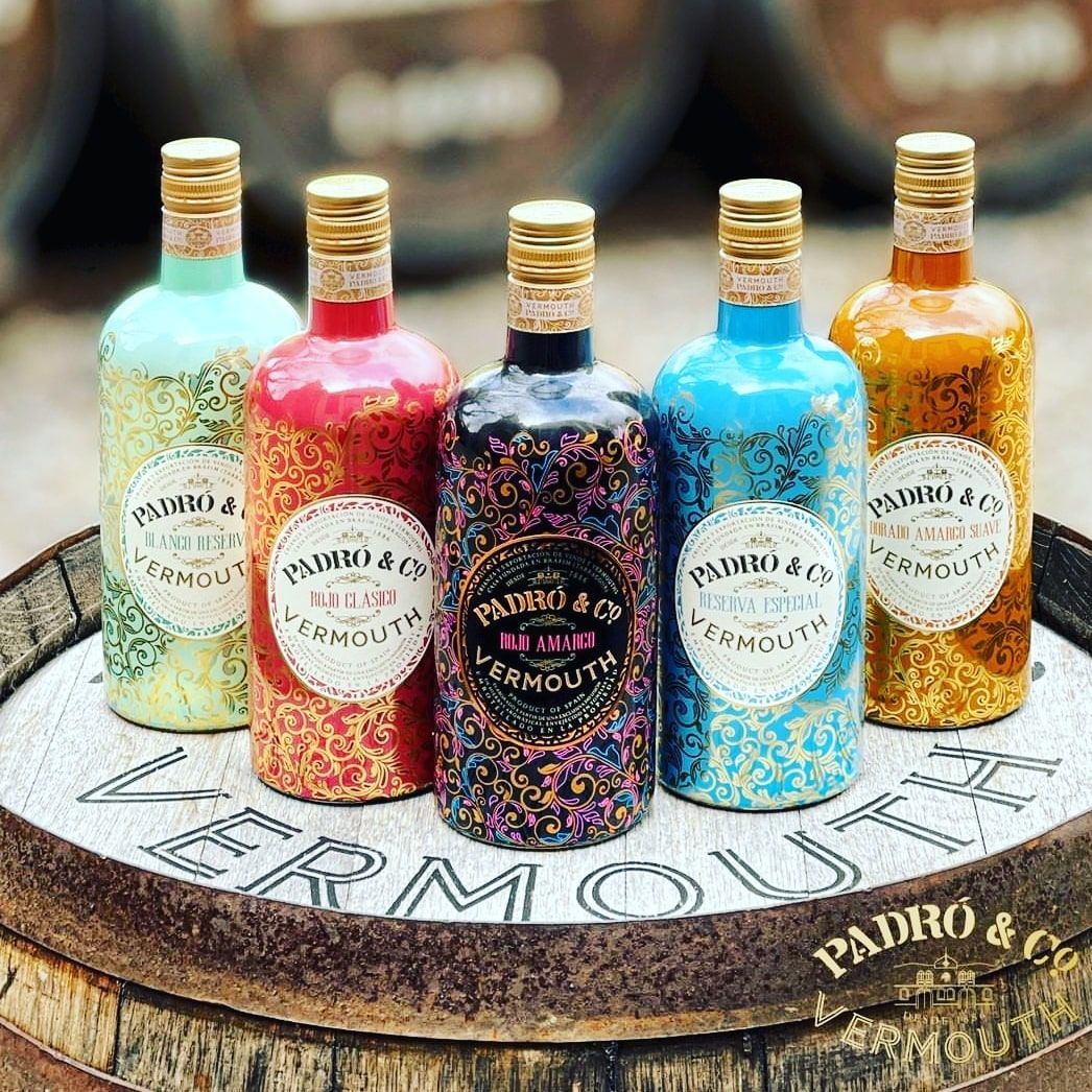 Cinco botellas de vermut Padró & Co., coloridas y con diseños variados, dispuestas sobre un barril de madera.