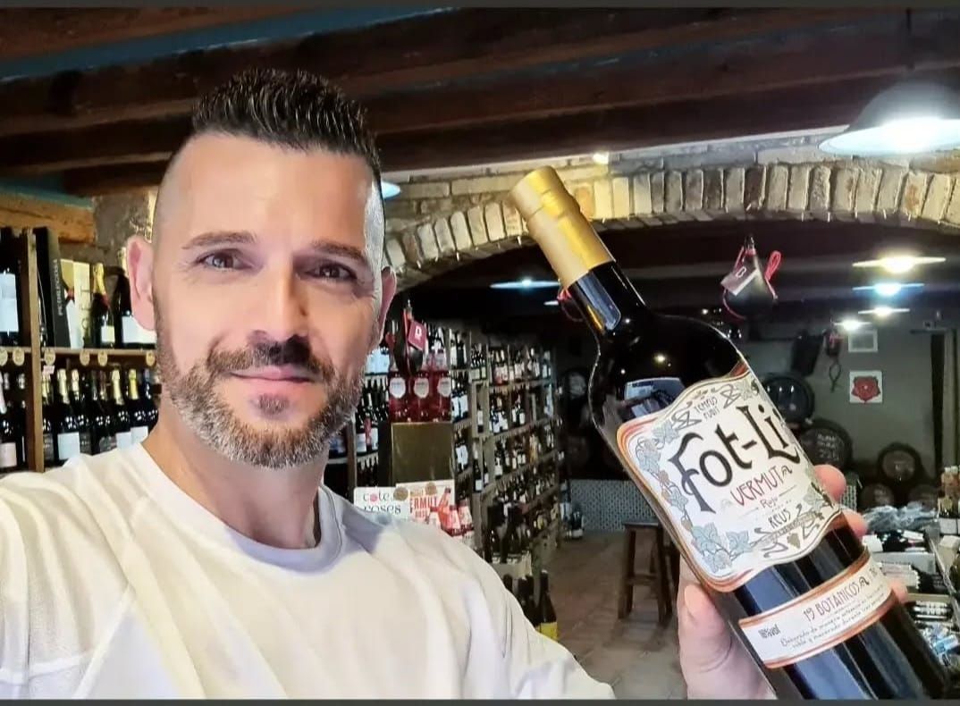 Una persona sonriente sostiene una botella de vermut Fot-Li en una vinoteca, con estanterías llenas de botellas al fondo.
