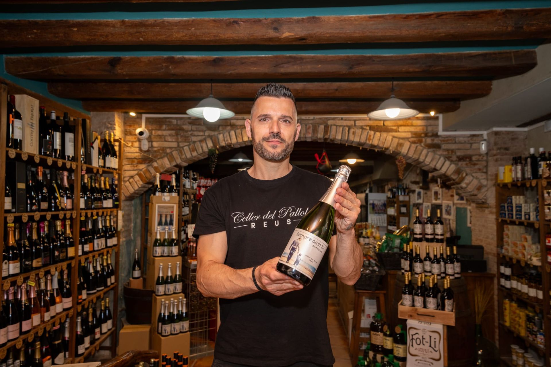 Un hombre se encuentra en una bodega, sonriendo mientras sostiene una botella de vino espumoso para la cámara.