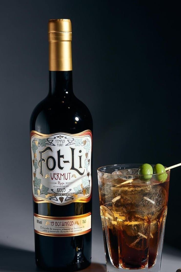 Una botella de vermut Fot-Li junto a un vaso lleno de hielo, un líquido oscuro y dos aceitunas ensartadas en un palillo.