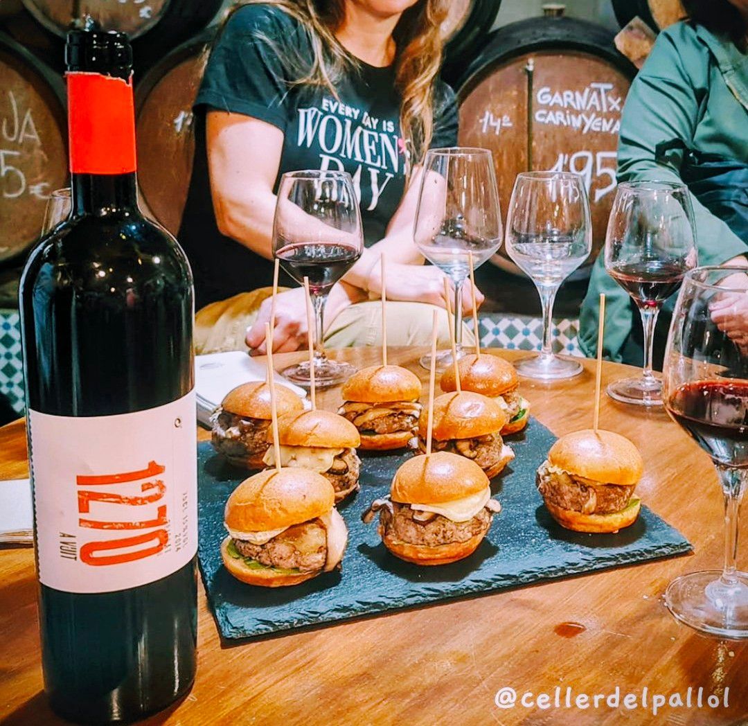 Una botella de vino y una bandeja de pizarra con seis mini hamburguesas sobre una mesa, con gente y copas de vino al fondo.