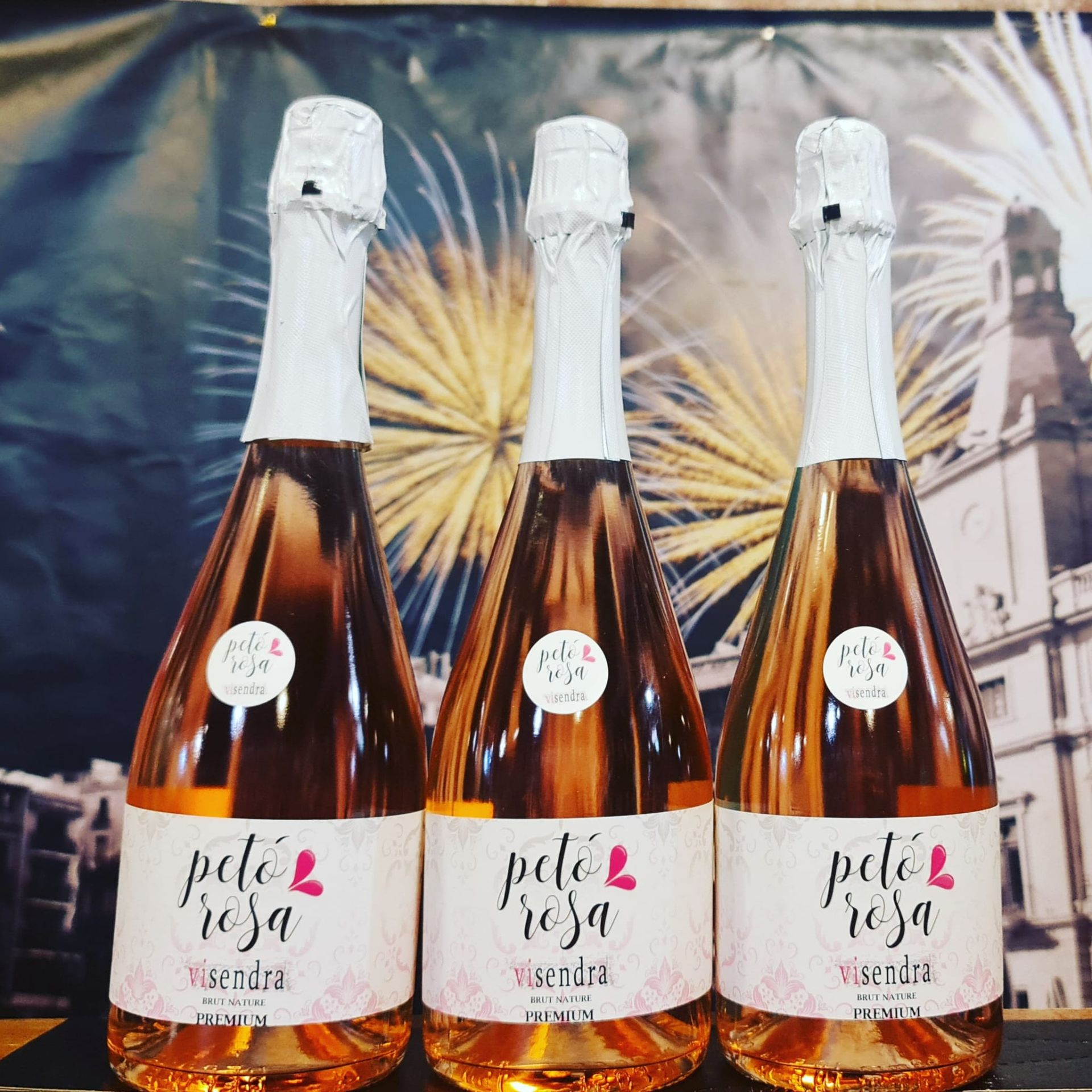 Tres botellas de vino rosado espumoso Petó Rosa se alinean con un telón de fondo de fuegos artificiales