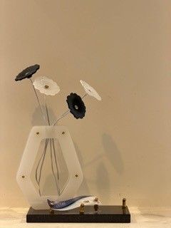 Un vase orné de fleurs en noir et blanc repose sur un support en bois. Un petit poisson en verre est placé en dessous.