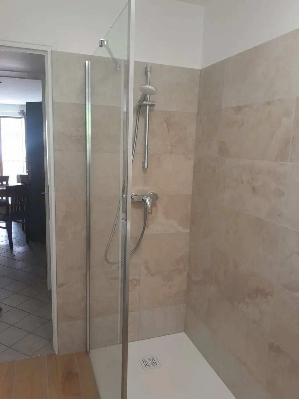 Douche avec porte vitrée, murs carrelés beiges et accessoires de douche argentés.