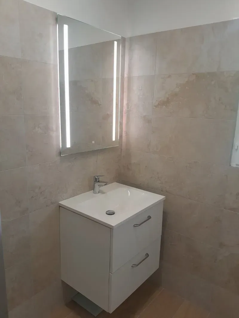 Coiffeuse blanche avec miroir, éclairée par des lumières verticales, contre des murs carrelés beiges.