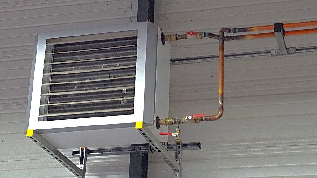 Un aérotherme industriel monté au plafond avec tuyauterie et vannes en cuivre, fixé à un support métallique.