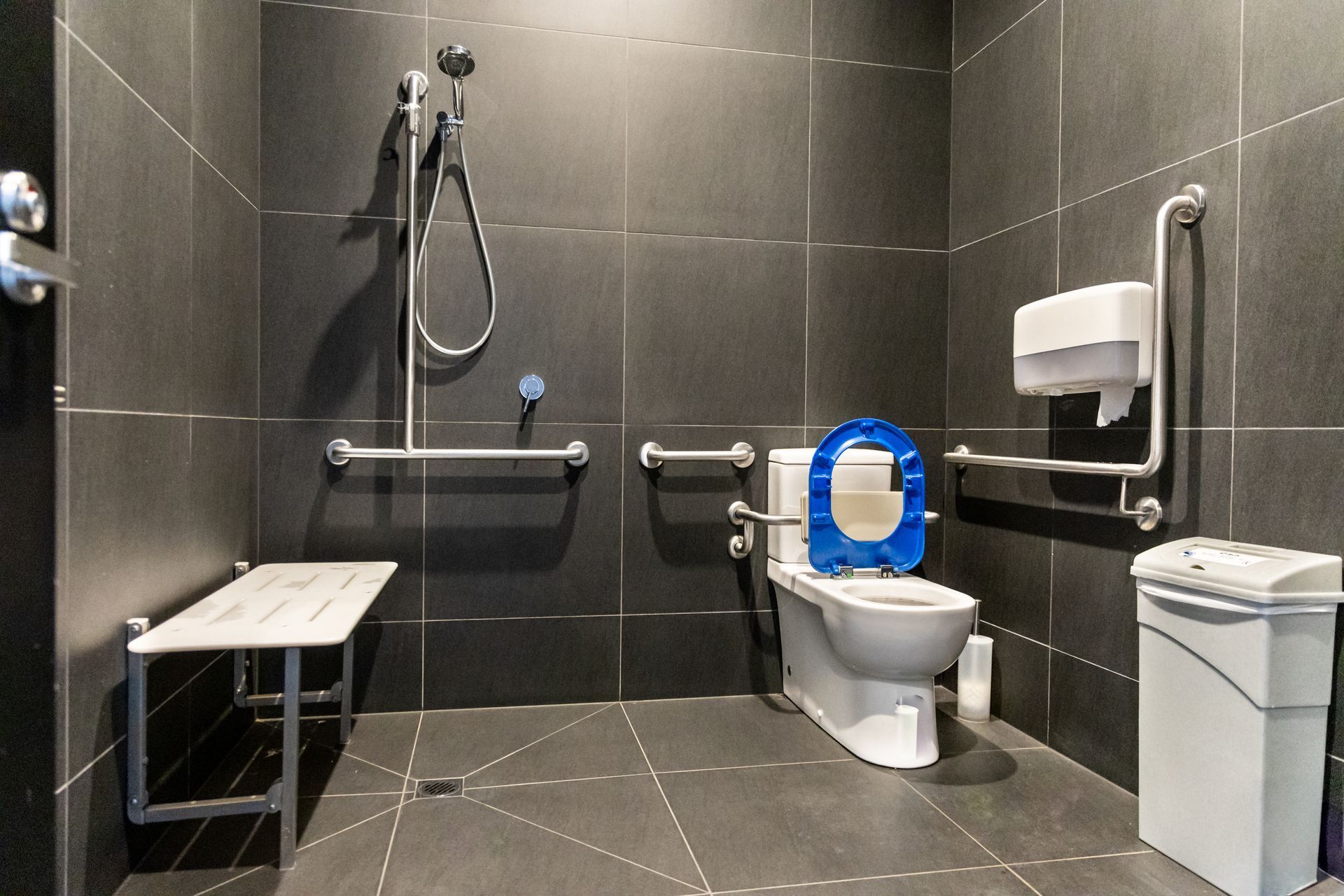 Salle de bains accessible aux PMR avec douche, toilettes, barres d'appui et banc rabattable ; carrelage gris foncé.