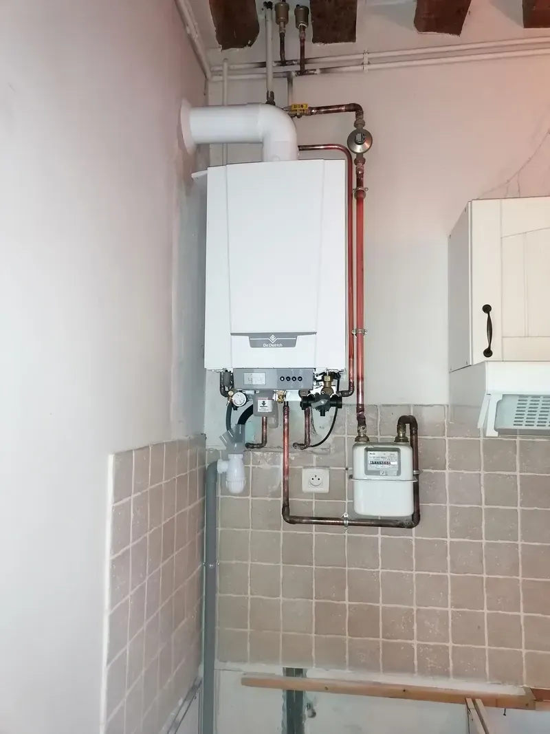 Chaudière à gaz murale blanche, tuyaux en cuivre, compteur de gaz et mur carrelé dans une cuisine.