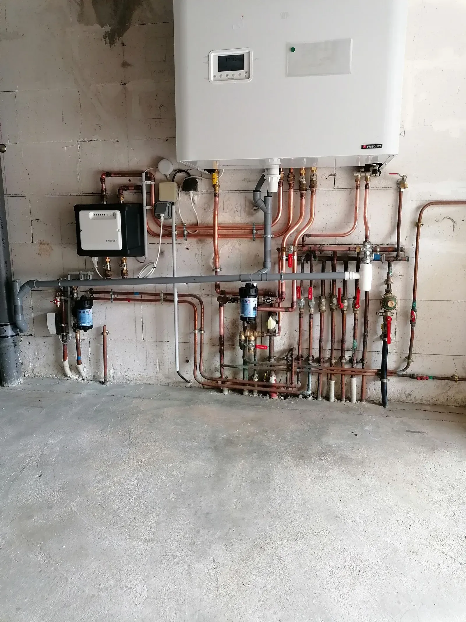 Tuyaux en cuivre et chaudière blanche fixés sur un mur en béton. Diverses vannes et raccords sont également présents.