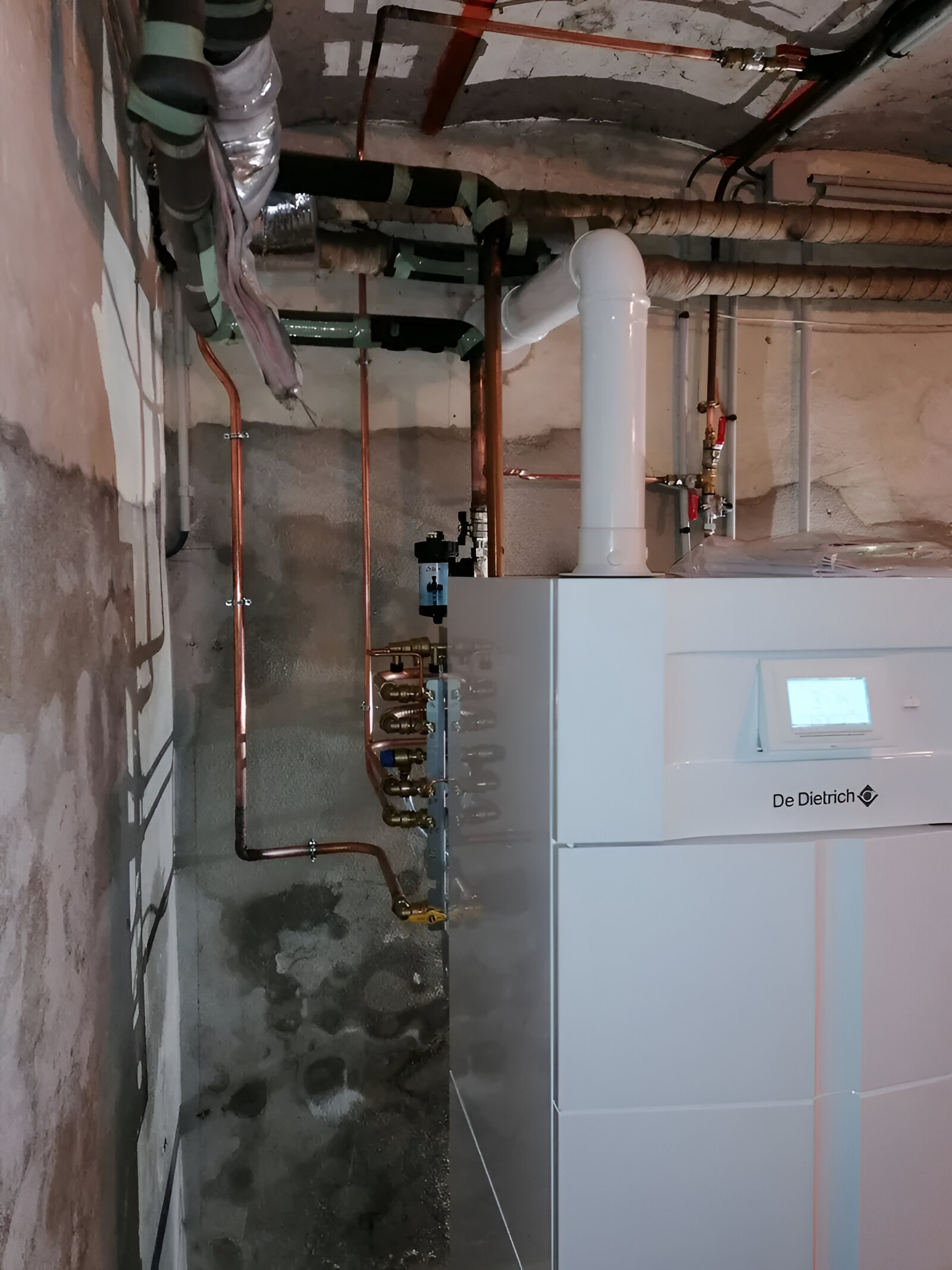 Un appareil de chauffage blanc avec des tuyaux en cuivre dans un sous-sol. Murs et plafond en béton gris.