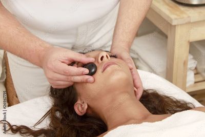 Eine Spa-Therapeutin, die einem liegenden Kunden auf einer Behandlungsliege eine Gesichtsmassage mit Steinen gibt