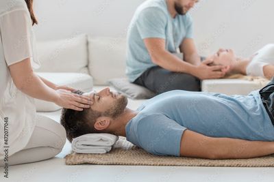 Personen, die in einem hellen Spa-Raum eine Reiki-Massage erhalten: Eine Person liegt und eine andere sitzt in der Nähe