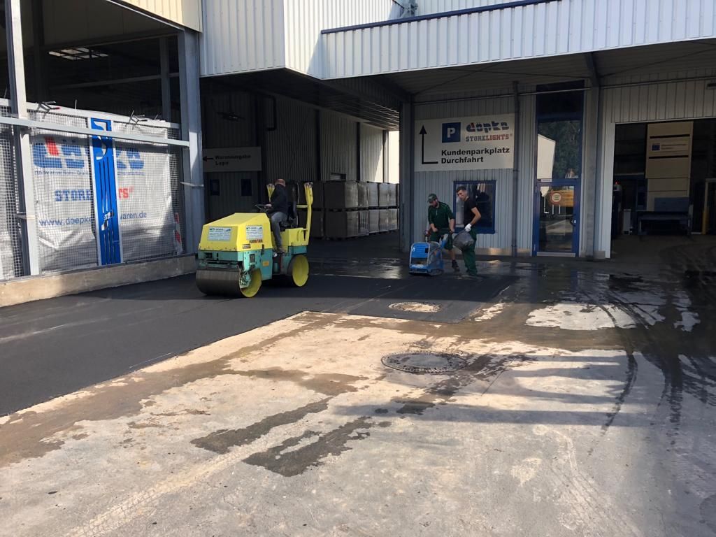 Eine Walze walzt Asphalt auf einem Parkplatz vor einem Gebäude.